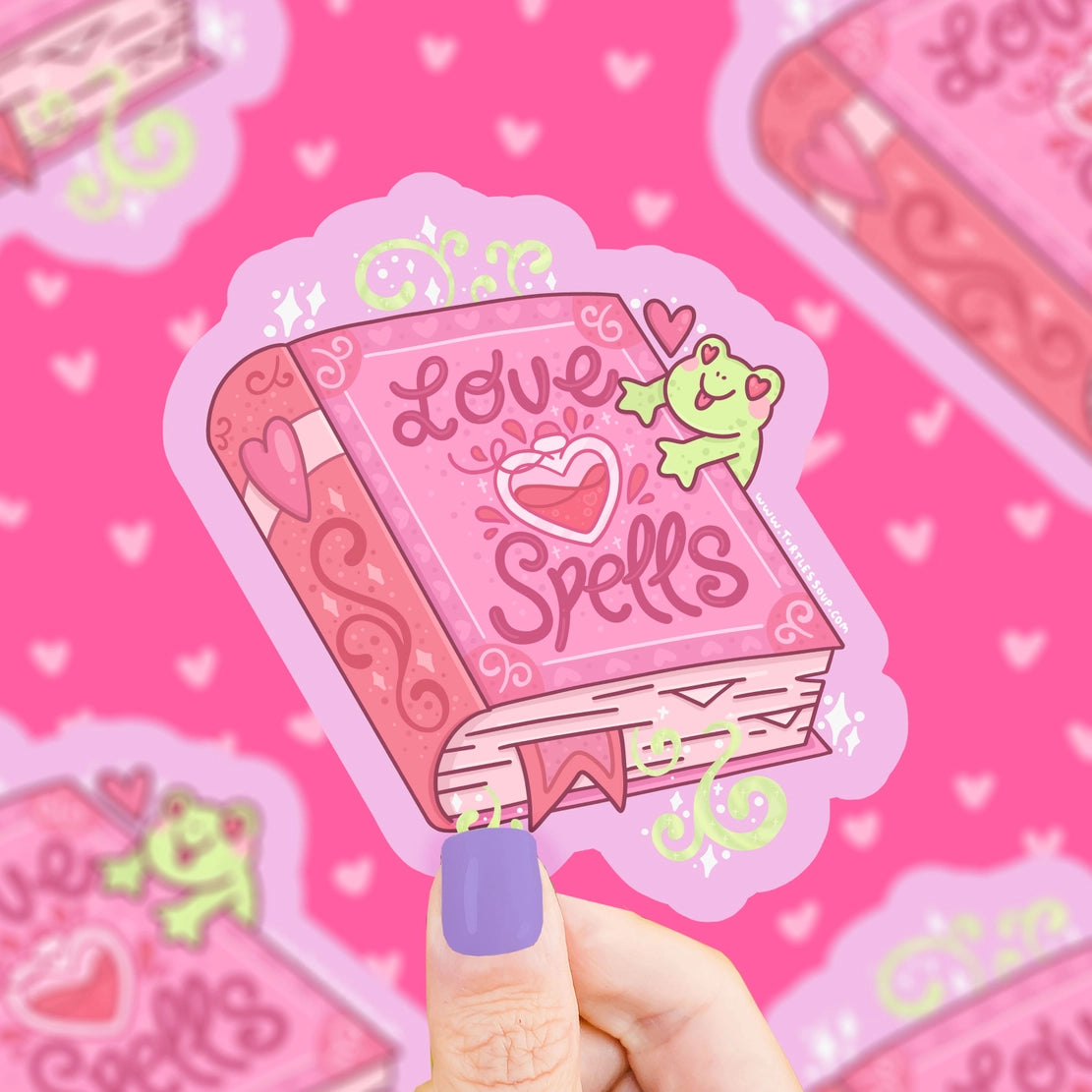 Hand holding a vinyl sticker of a pink love spells book with a smiling green frog and heart designs on a pink background