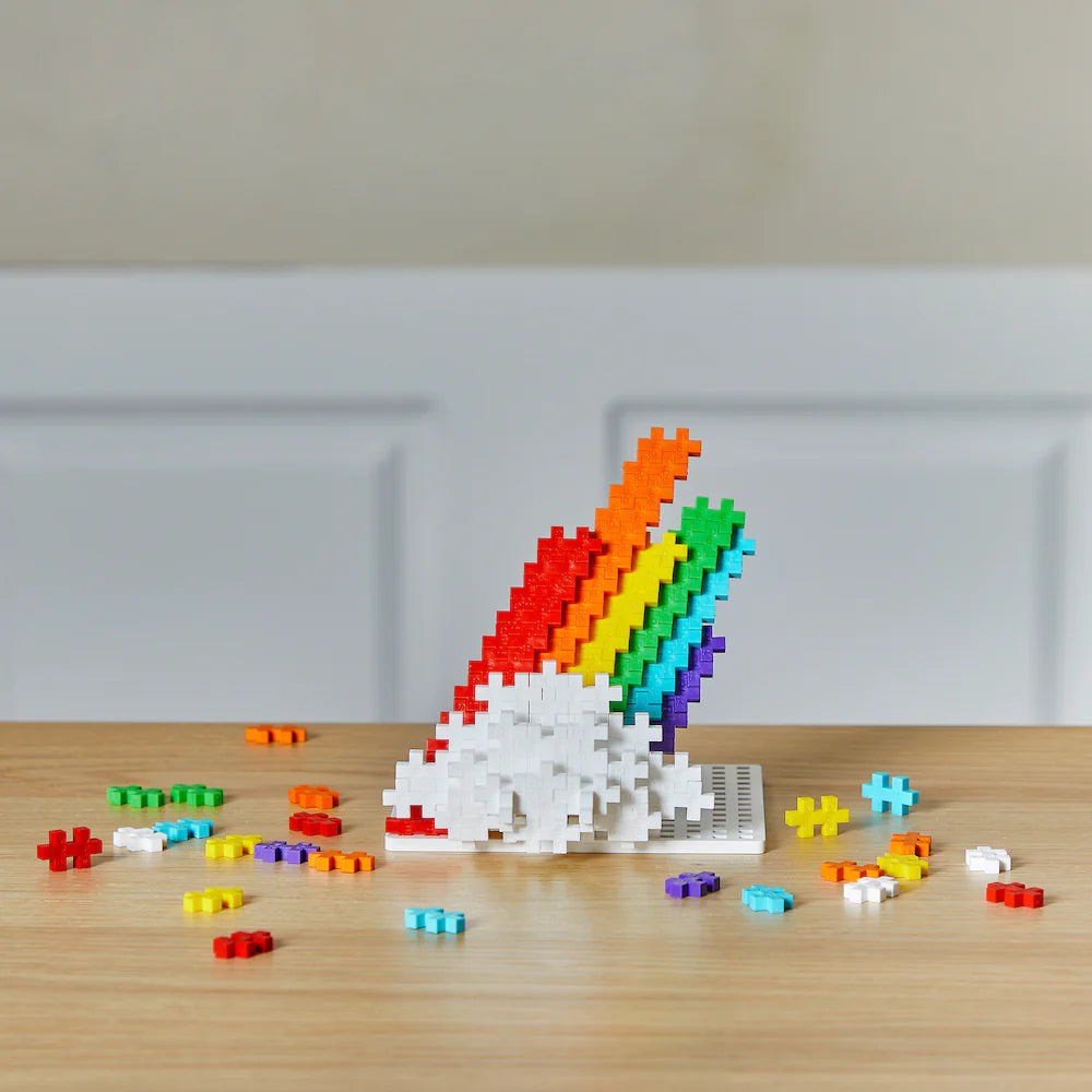 Plus Plus Rainbow 70pc features colorful building blocks arranged as a rainbow with white clouds on a wooden surface.