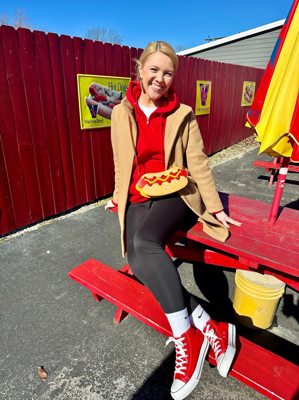 Handbag | Classic Hot Dog worn by a smiling woman sitting on a red bench outside with a bright sunny sky.