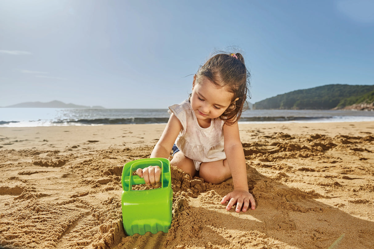 Little girl playing on sunny sandy beach using bright green toy shovel to dig in sand