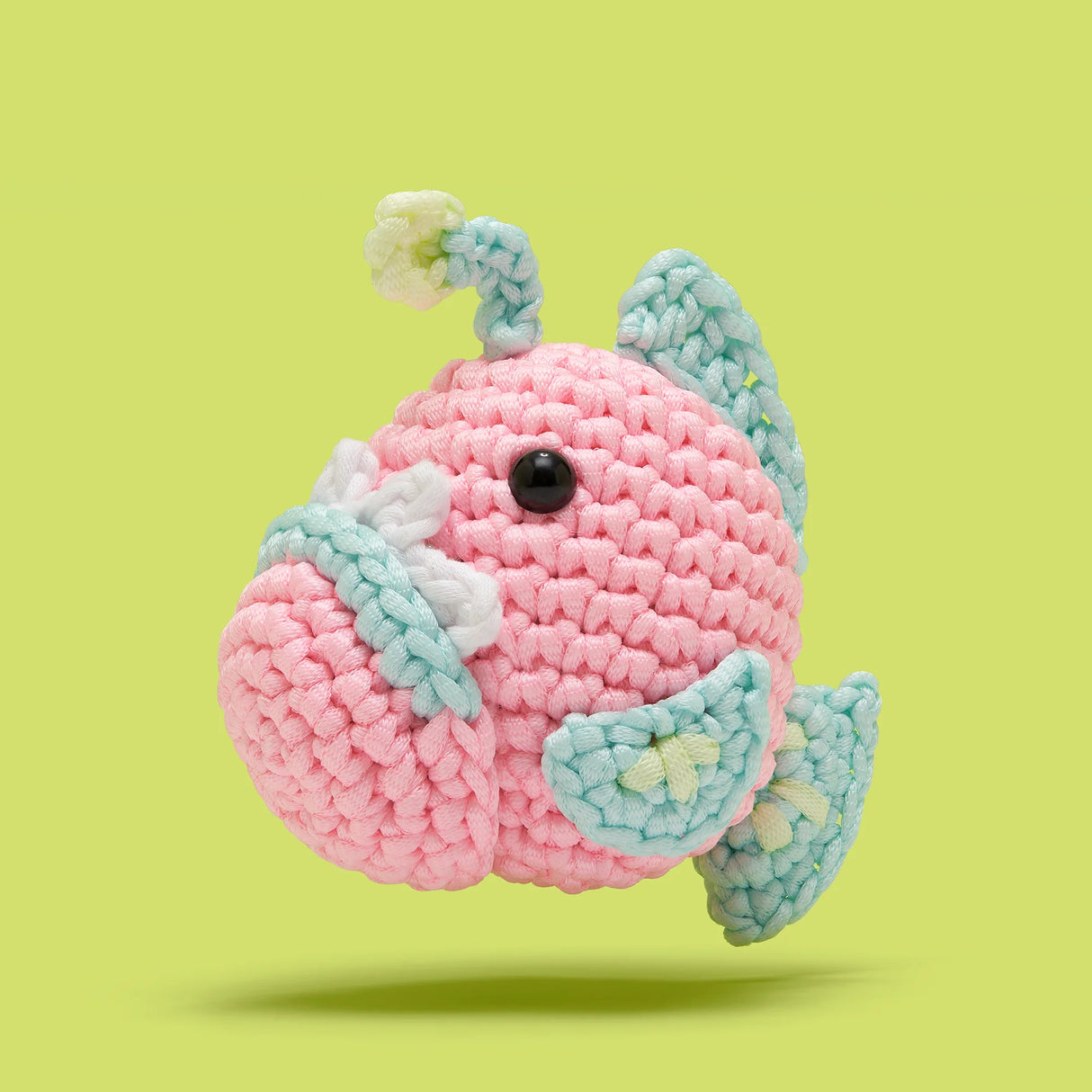 Crocheted pink and blue angler fish toy with large eyes and glowing lure on a light green background