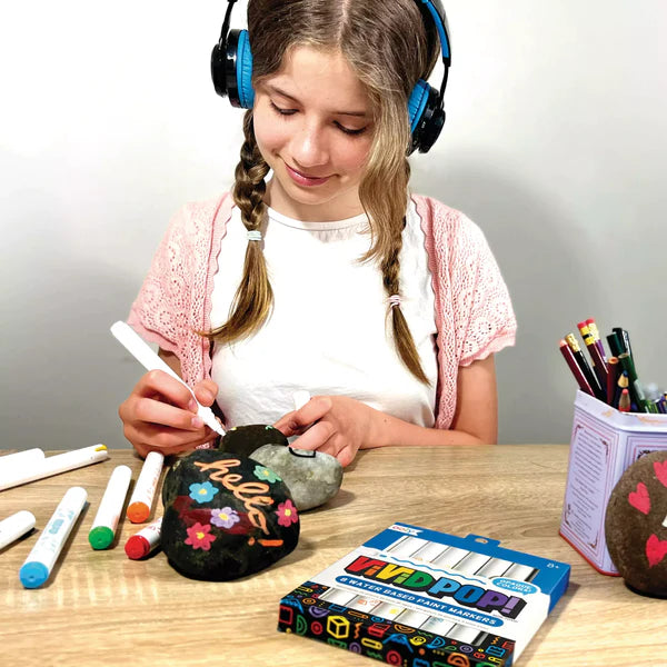 Girl painting rocks with colorful Vivid Pop! Paint Markers acrylic set on wooden table, creating vibrant art designs.