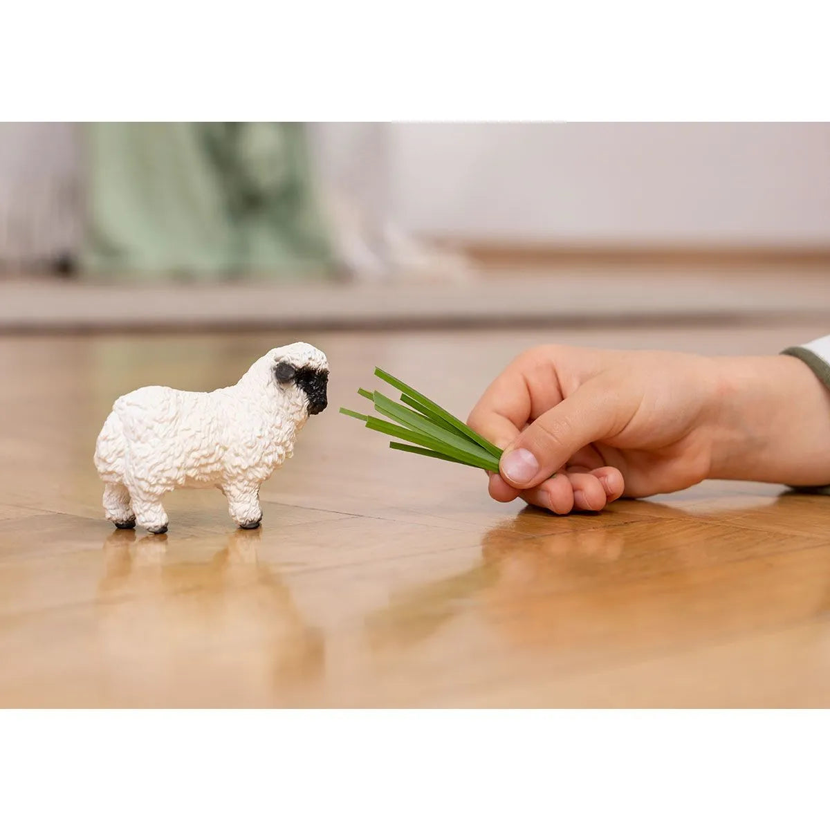 Valais Black-Nosed Sheep toy with white wool and black face next to a child's hand offering green grass on wooden floor.
