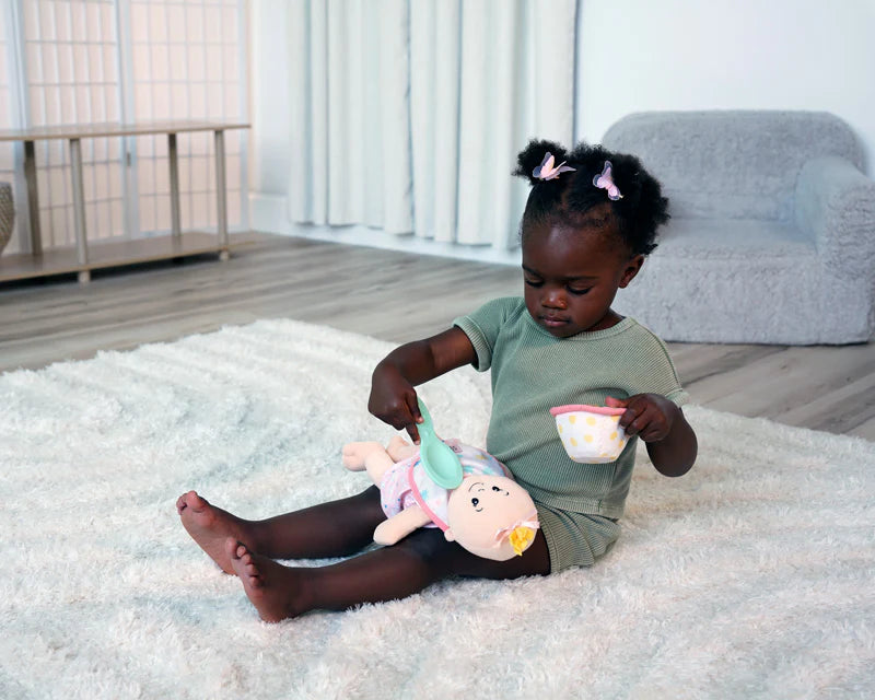Toddler girl in green plays on white rug feeding soft doll with pastel pink and yellow cup and spoon