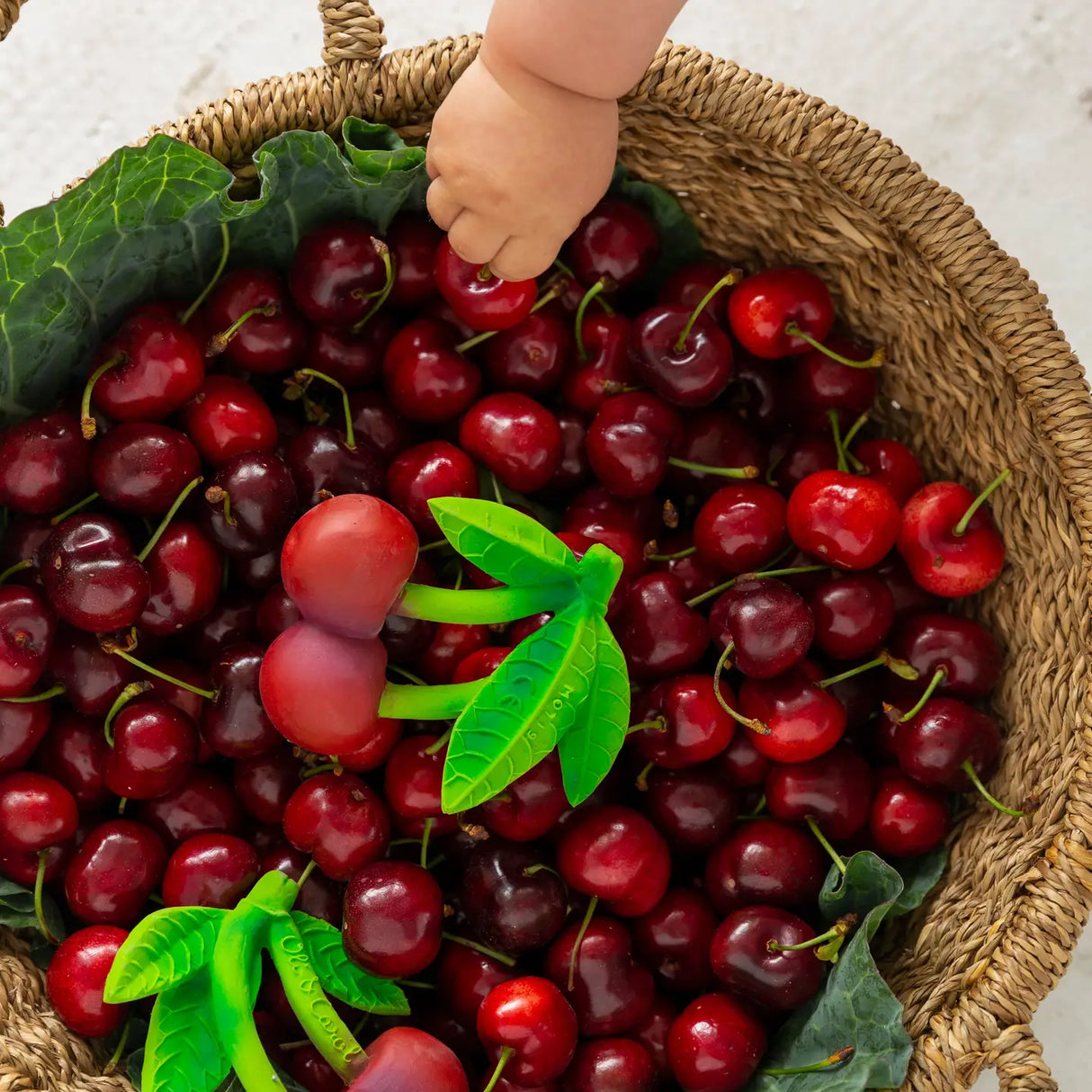 Baby’s hand reaching for a natural cherry-shaped teether among fresh cherries in a woven basket with green leaves