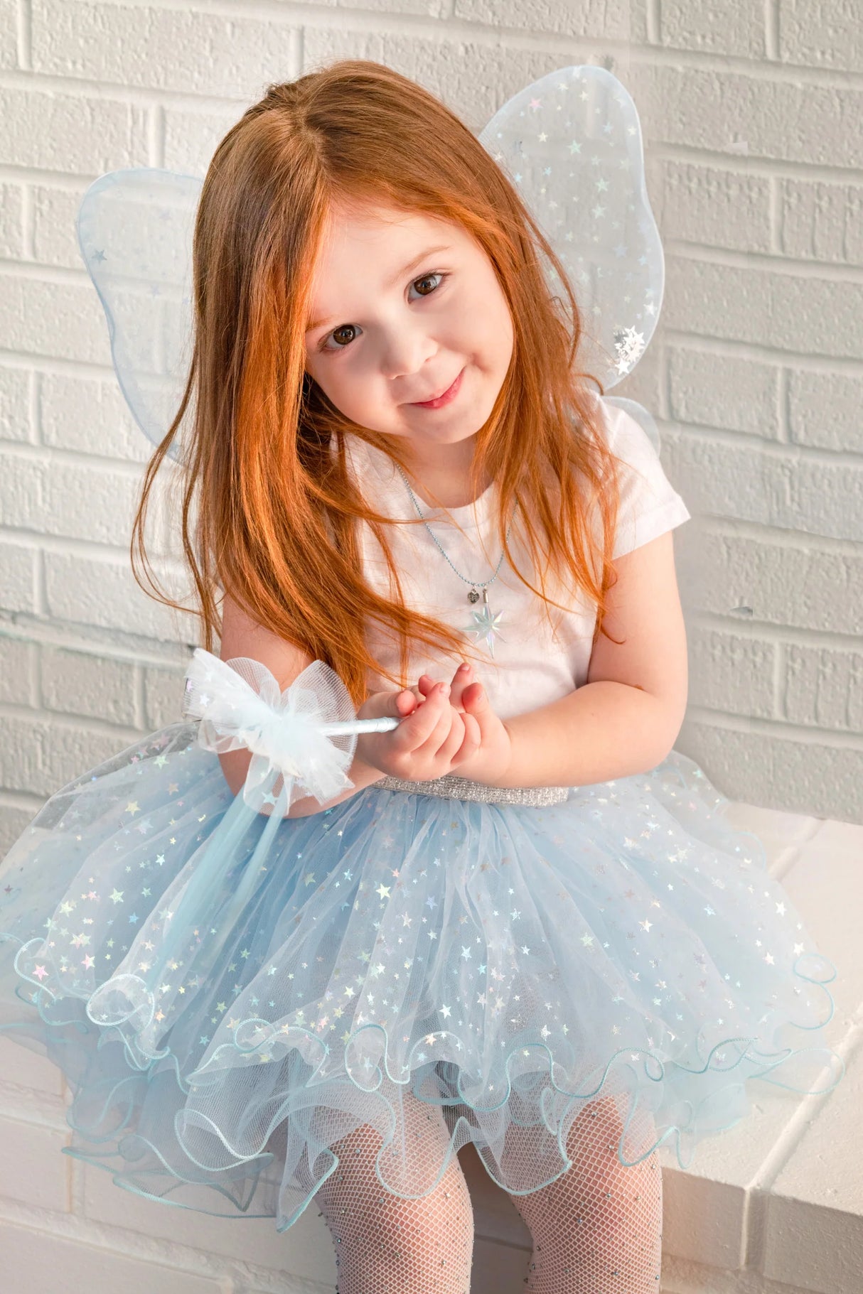 Young girl wearing light blue fairy wings and tutu with silver stars holding a matching star wand sitting by white brick wall.
