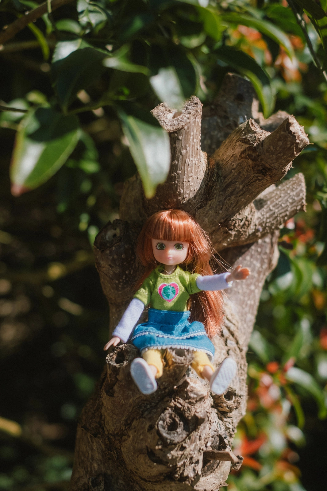 Lottie Planet Rescuer doll with green shirt and denim skirt sitting on tree branch outdoors surrounded by greenery