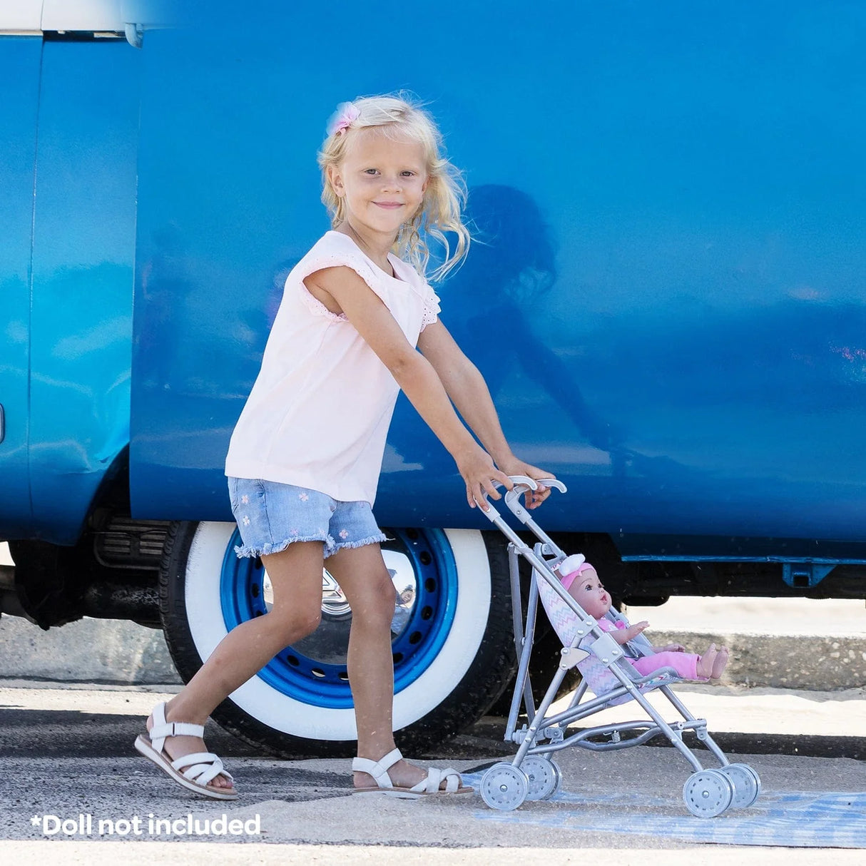 Little girl pushing a doll in the Adora Mini Stroller with Stroller Umbrella Zig Zag Rainbow outside near a blue vehicle.