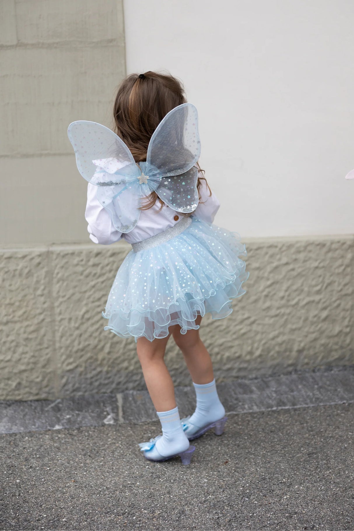 Child wearing light blue tutu and shimmering fairy wings with silver star details outdoors on pavement