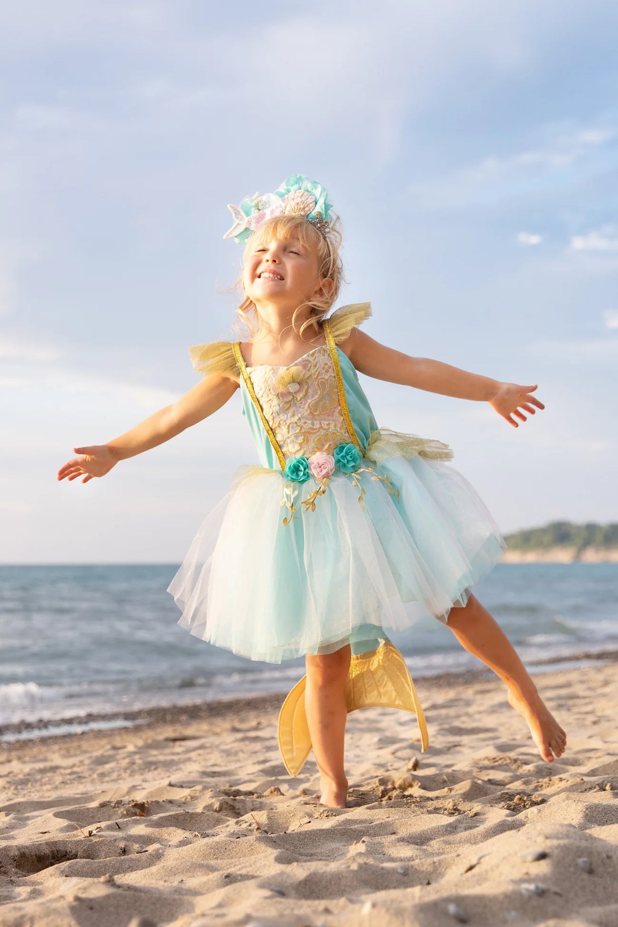 Young girl joyfully wearing the Mermalicious Dress with Tail at the beach, featuring a shimmery sea blue skirt and floral details.