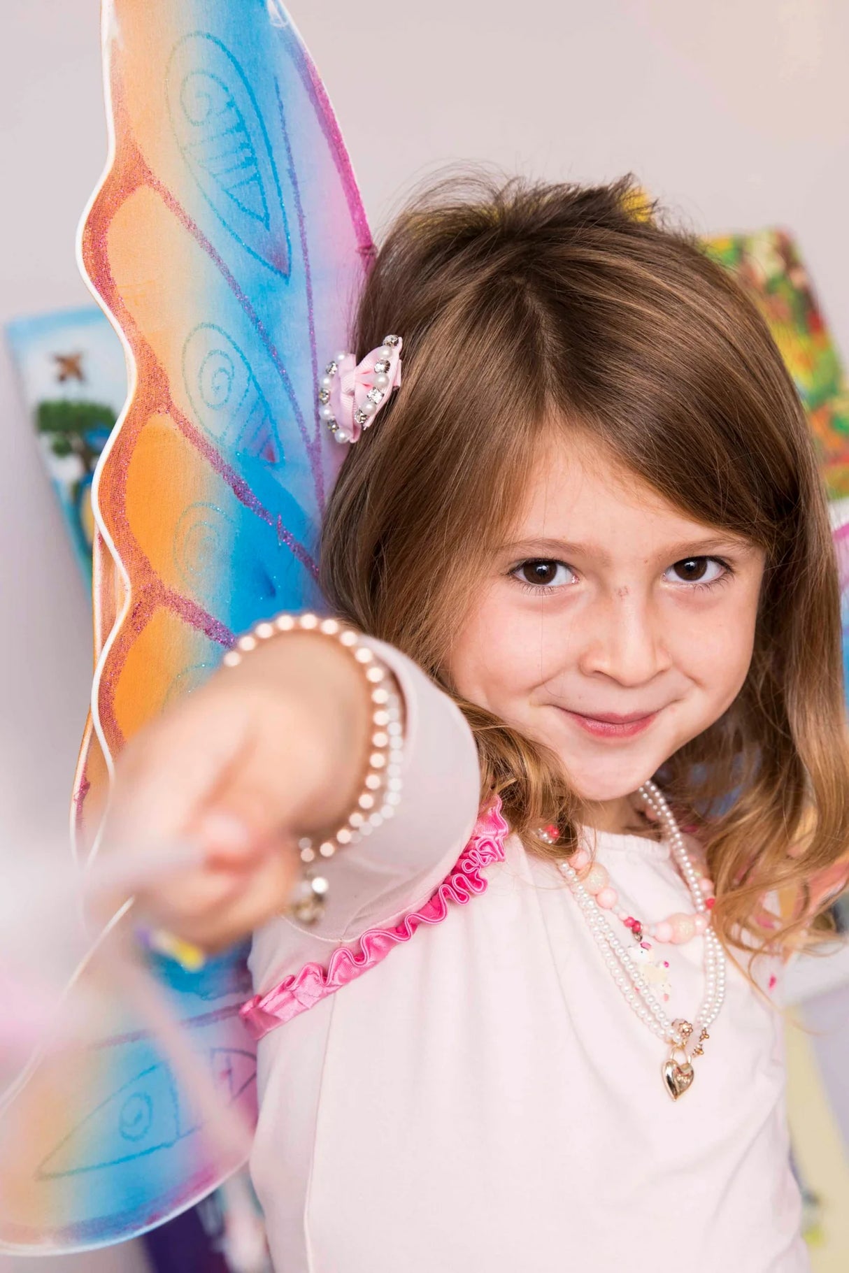 Smiling child wearing colorful Glitter Rainbow Wings with pink accents and pearl jewelry, playing indoors.