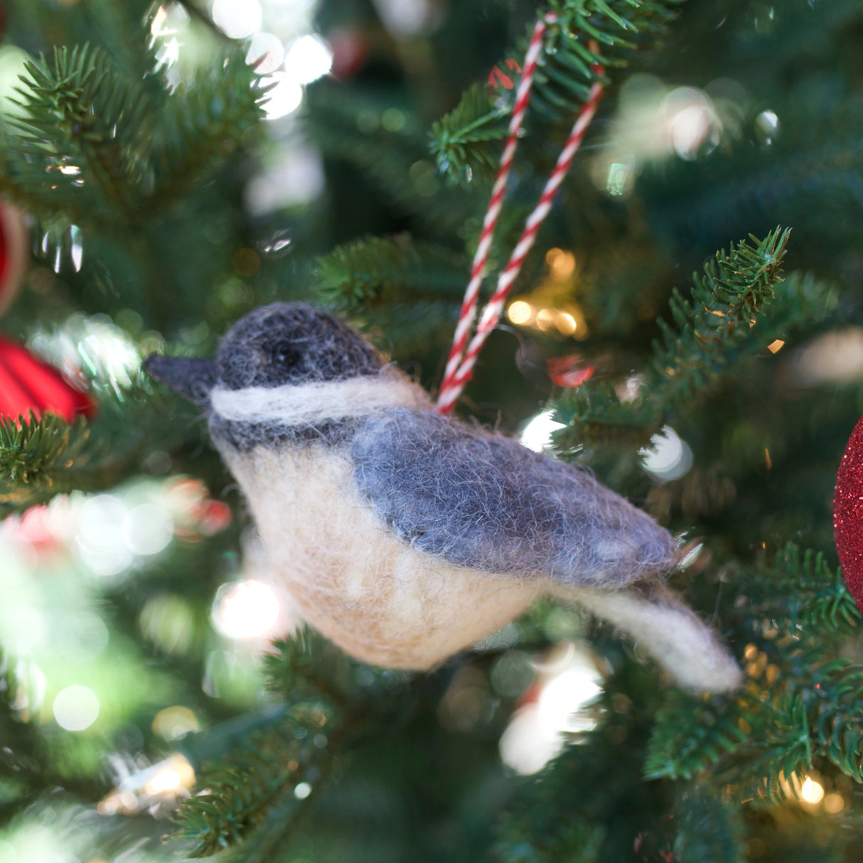 Handmade felt wool bird ornament in gray and cream hanging on a Christmas tree branch with lights nearby