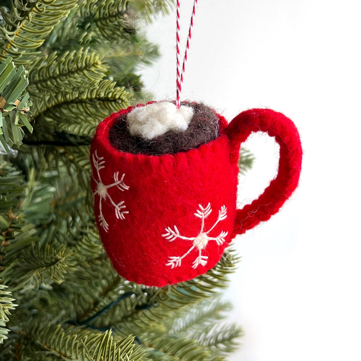 Handmade red felt ornament shaped like a hot chocolate mug with snowflake design and marshmallows hanging on a Christmas tree branch
