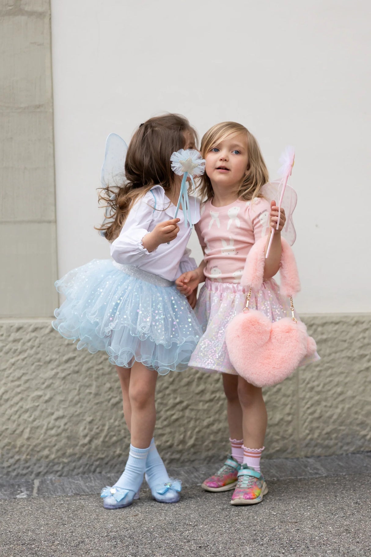 Two young girls wearing fairy wings, tutus, and holding matching wands, sharing a playful moment outdoors.
