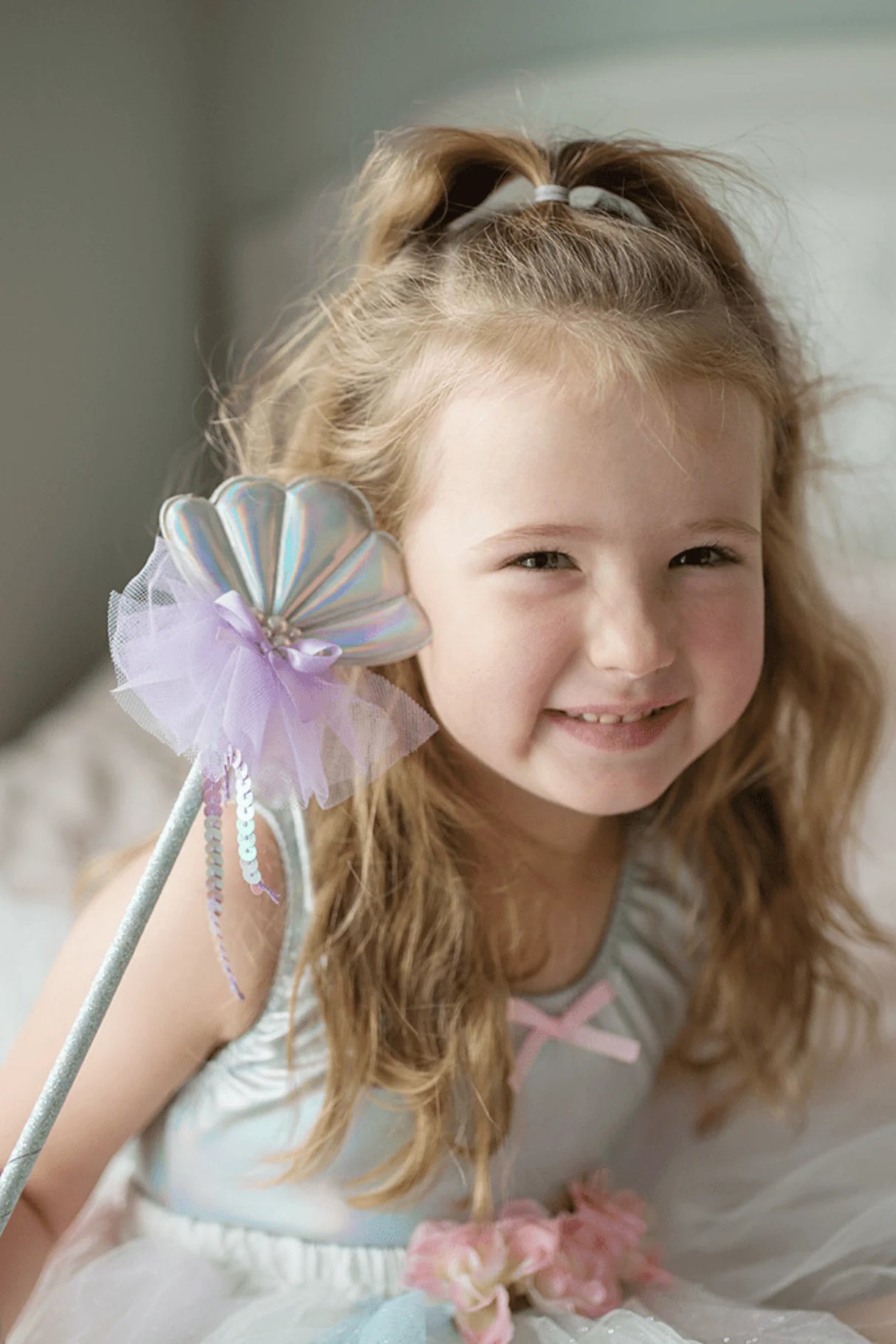 Young girl smiling and holding a Mermaid Wand with soft tulle and glittery details in light pastel colors.
