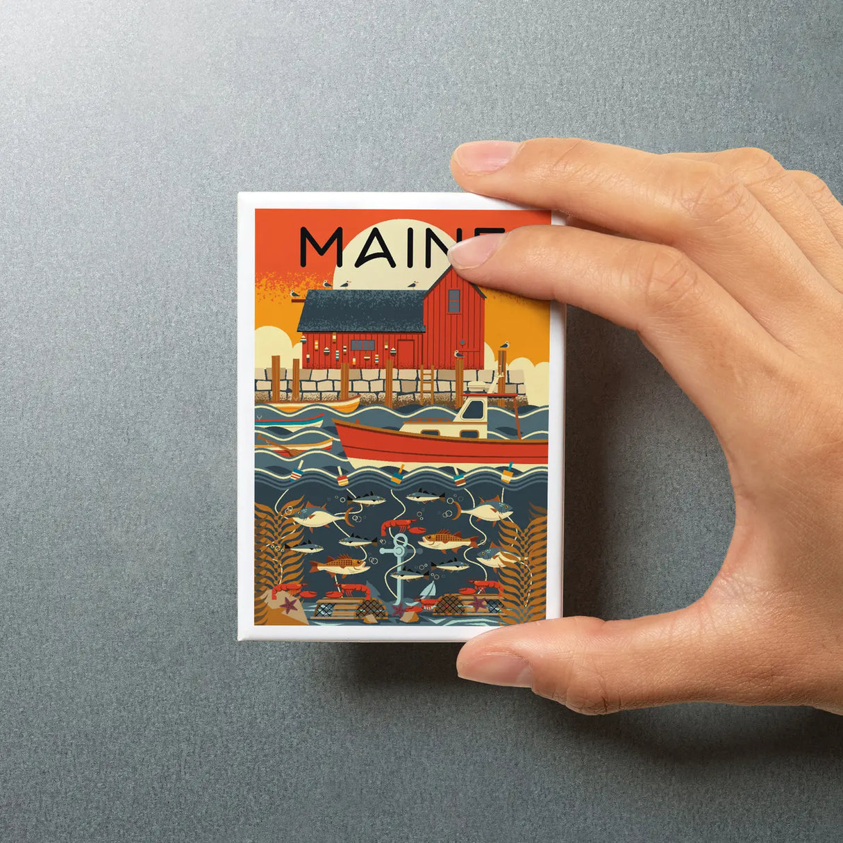 Hand holding a Maine Nautical Geometric Magnet featuring a colorful coastal scene with a boat and fish on a silver background.
