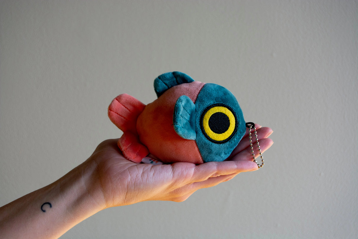Hand holding a soft, round mochi fish fry plush keychain with vibrant blue and orange colors and a small chain attached