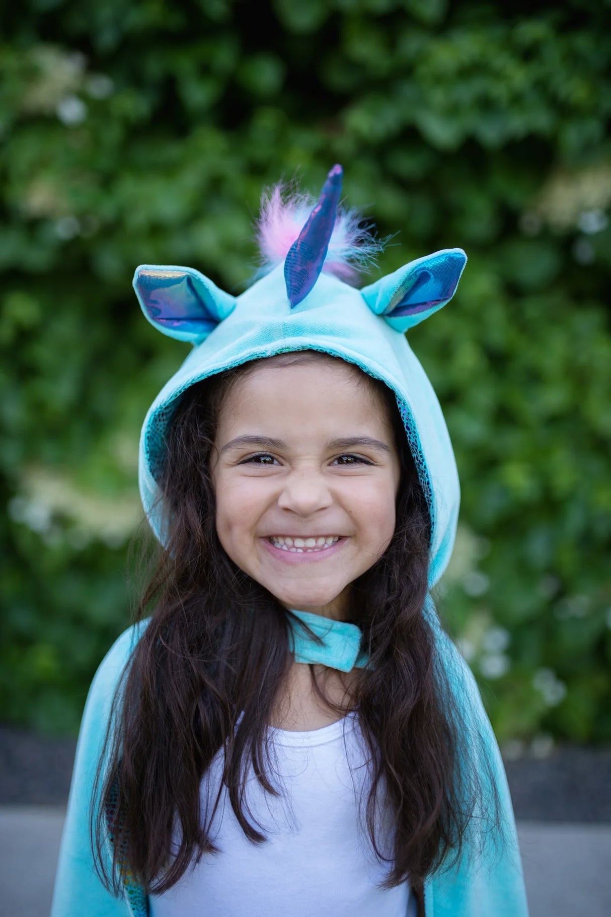Smiling child wearing the Unicorn Dragon Reversible Cape with blue hood, unicorn horn, ears, and fluffy mane outdoors.