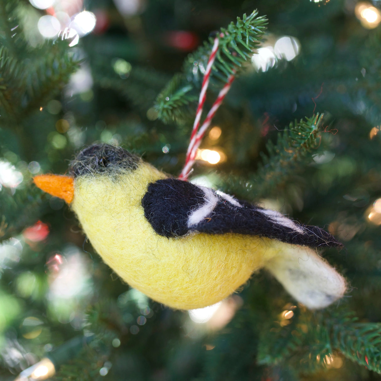 Handmade felt bird ornament in yellow and black wool hanging on a Christmas tree branch with festive lights.