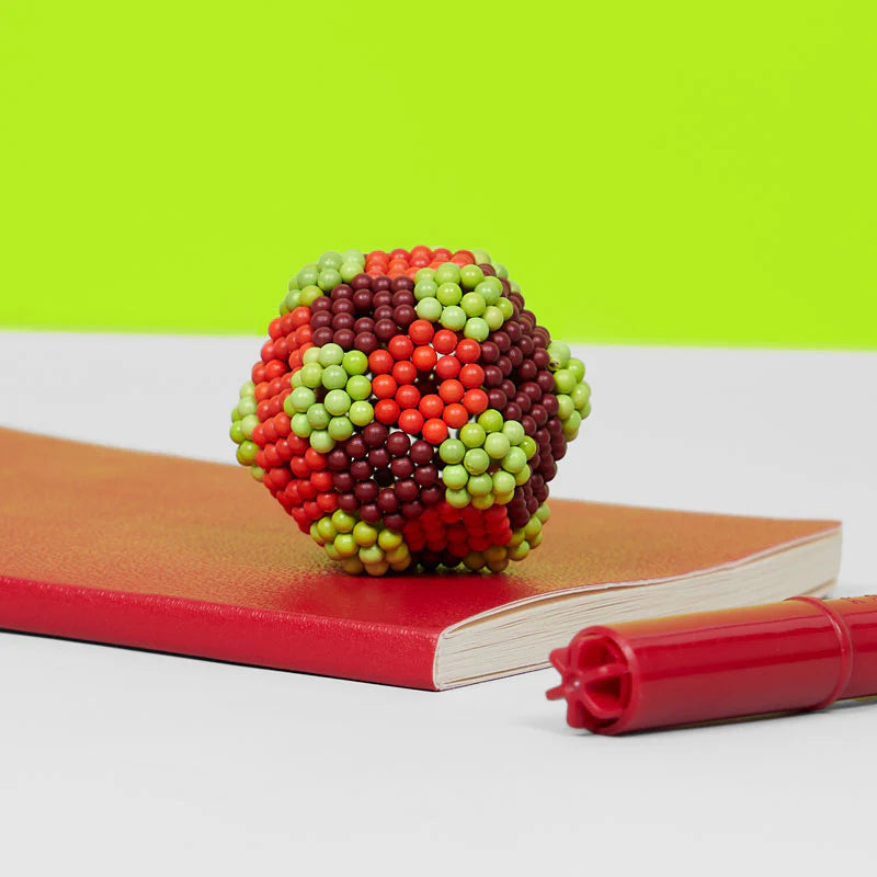 Speks. Bolds. Brash magnetic balls shaped into a colorful cluster on a red notebook with a pen nearby.