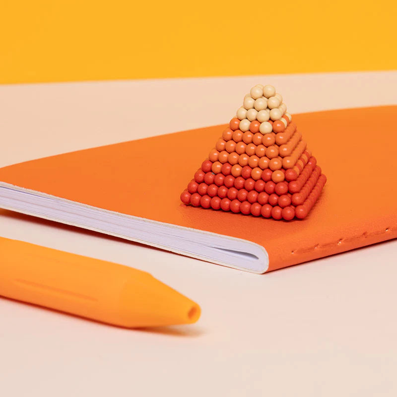 Speks. Pixel. 1 Up. colorful magnetic balls stacked into a pyramid on notebook with orange pen nearby.