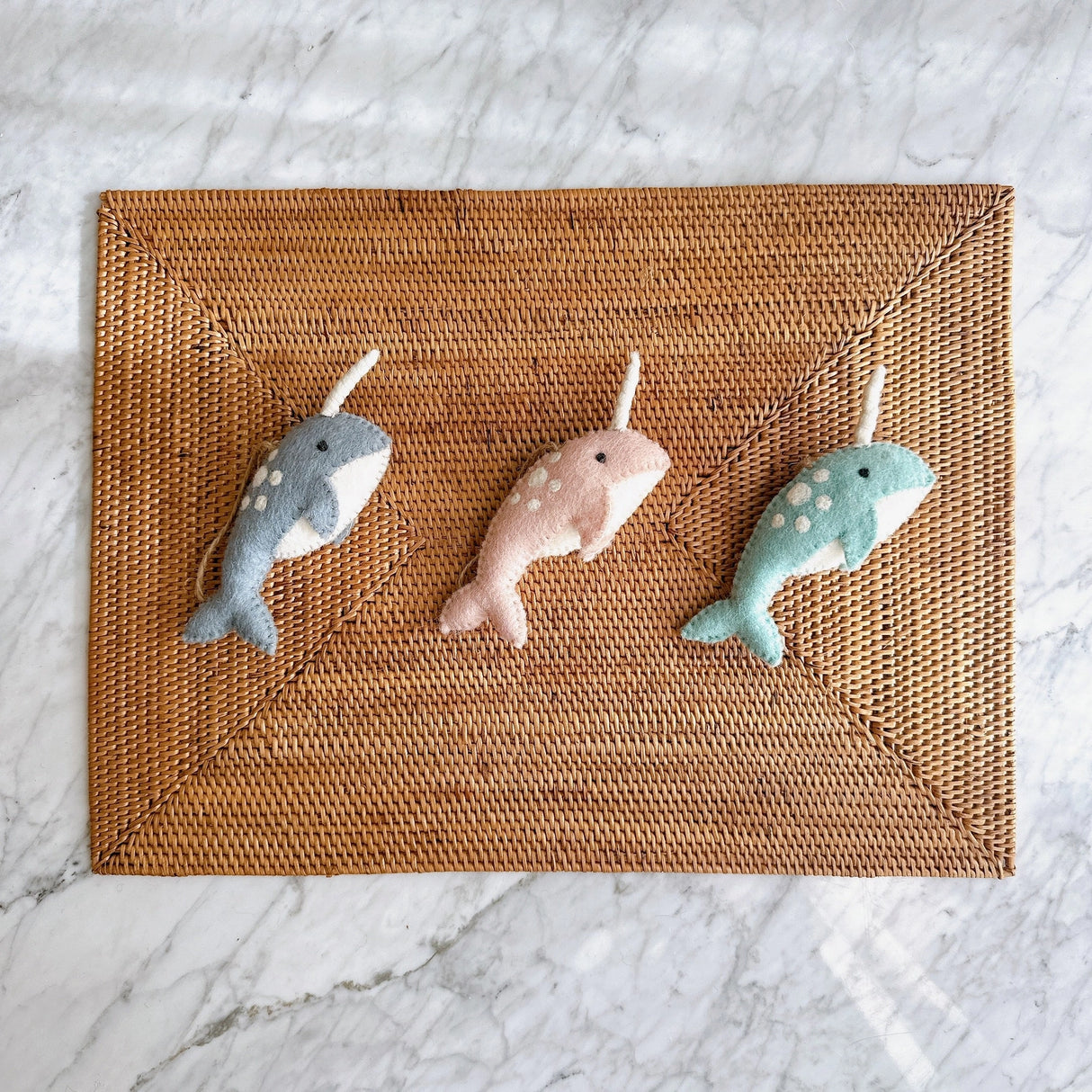 Three hand-felted narwhal ornaments in blue, pink, and teal on a woven placemat background