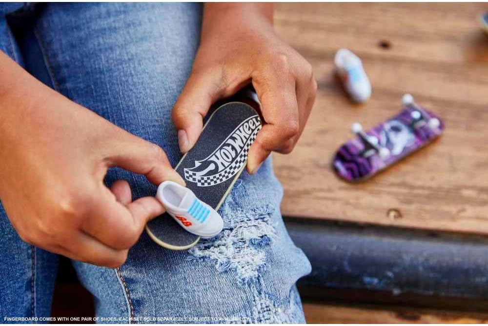 Close-up of hands holding Hot Wheels Skateboard & Shoes mini skateboard toy over ripped jeans, with additional shoes visible.