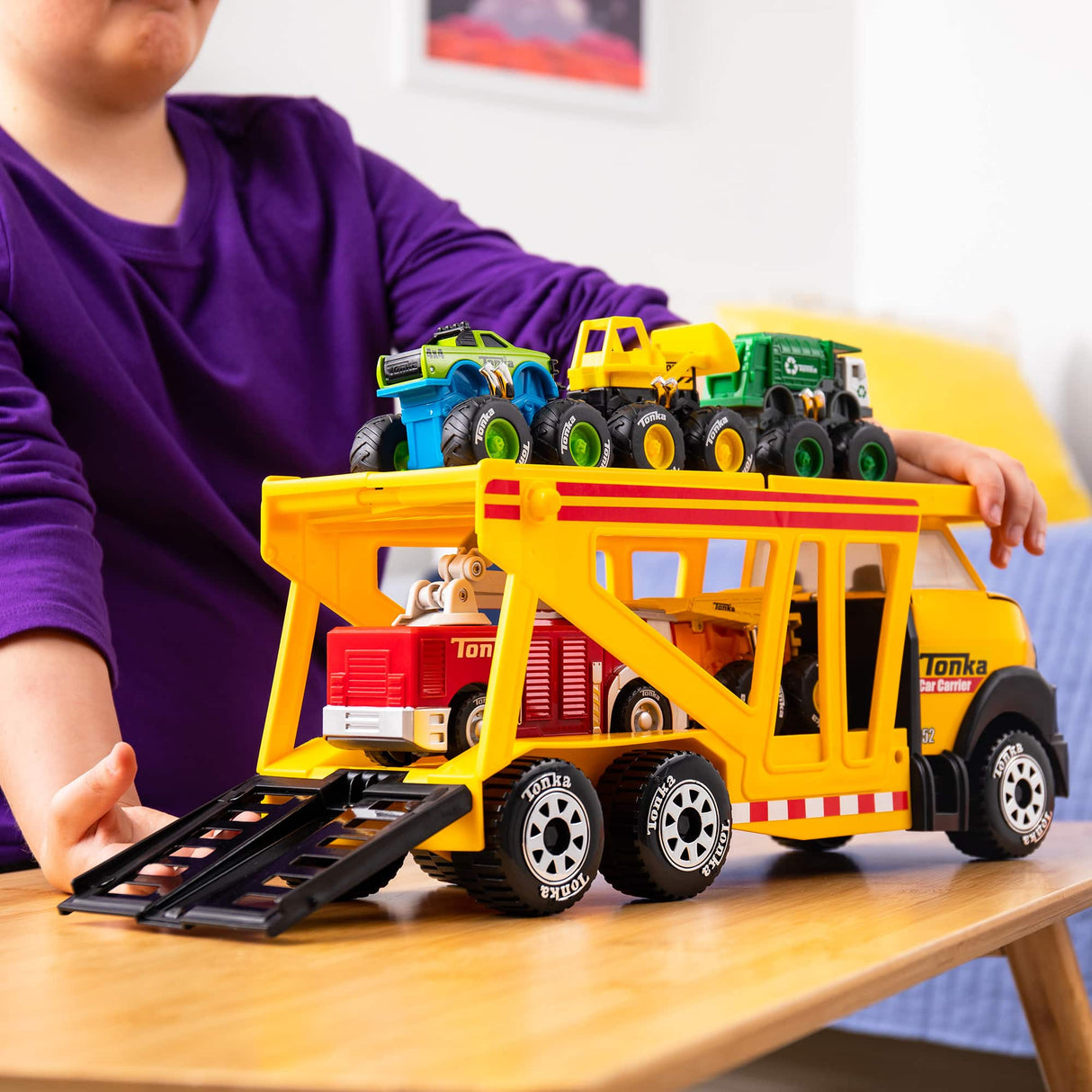 Child playing with Tonka Car Carrier toy truck loaded with multiple vehicles on a wooden table indoors.
