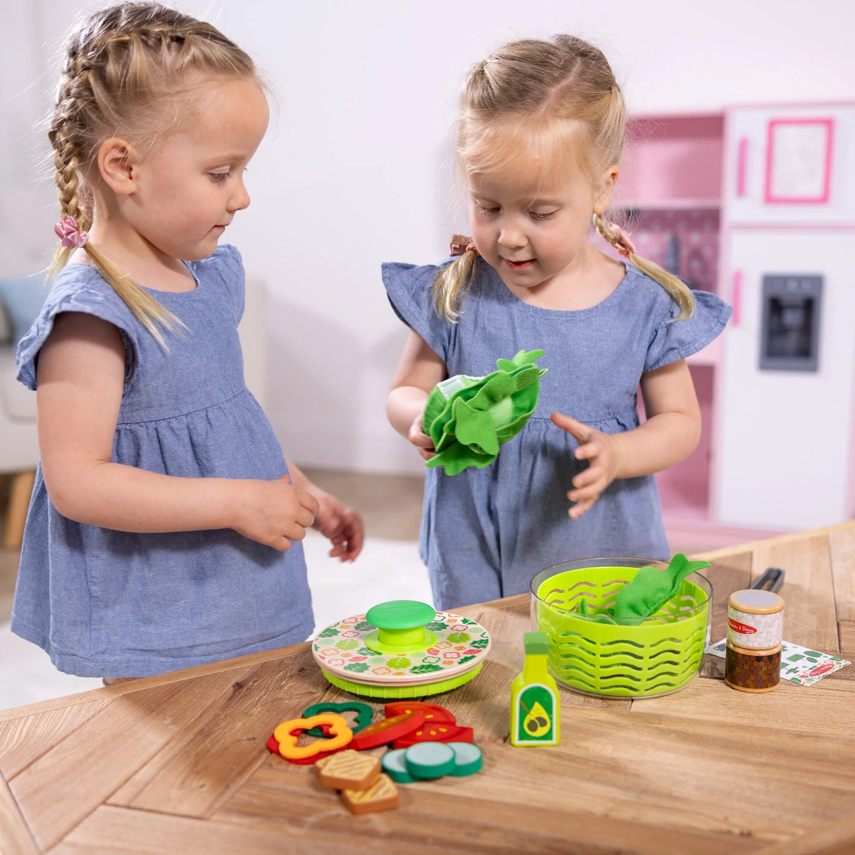 Two young girls playing with the Salad Spinner toy set featuring fabric lettuce and wooden vegetables on a table.