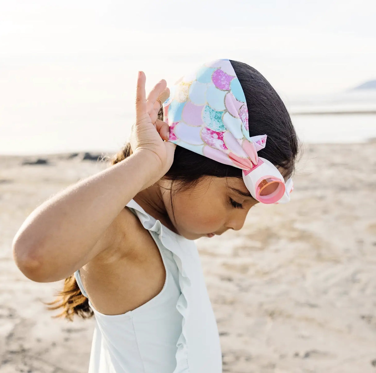 Child wearing pink and teal Goggles Mermaid with mermaid scale strap on sandy beach preparing to swim.