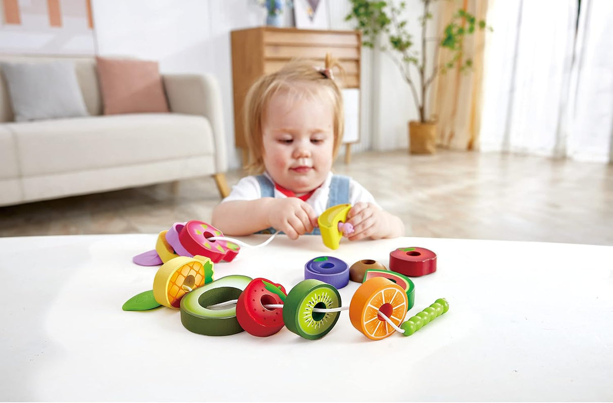 Toddler playing with colorful wooden Caterpillar Fruit Feast Set and threading fruit-shaped pieces on a string at a table