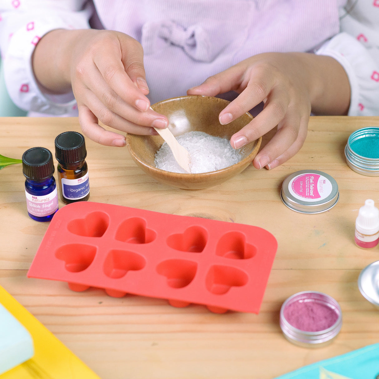 Hands mixing ingredients in a bowl with heart-shaped mold for DIY Bath Bombs Kit on a wooden table.