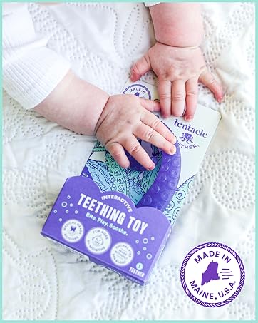 Baby holding purple Tentacle Teether interactive teething toy in packaging on white quilt.