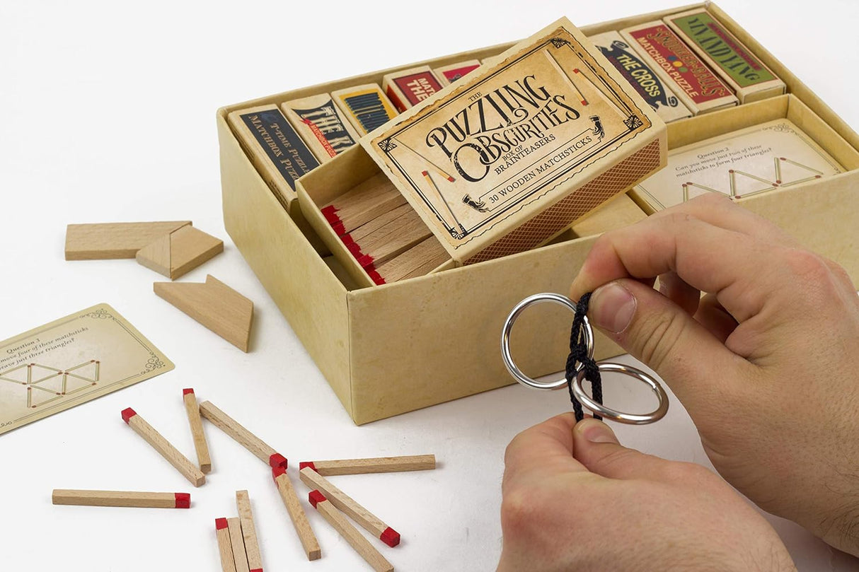 Hands solving metal ring puzzle next to wooden sticks and cards from the Puzzling Obsurities Matchbox Puzzle set.