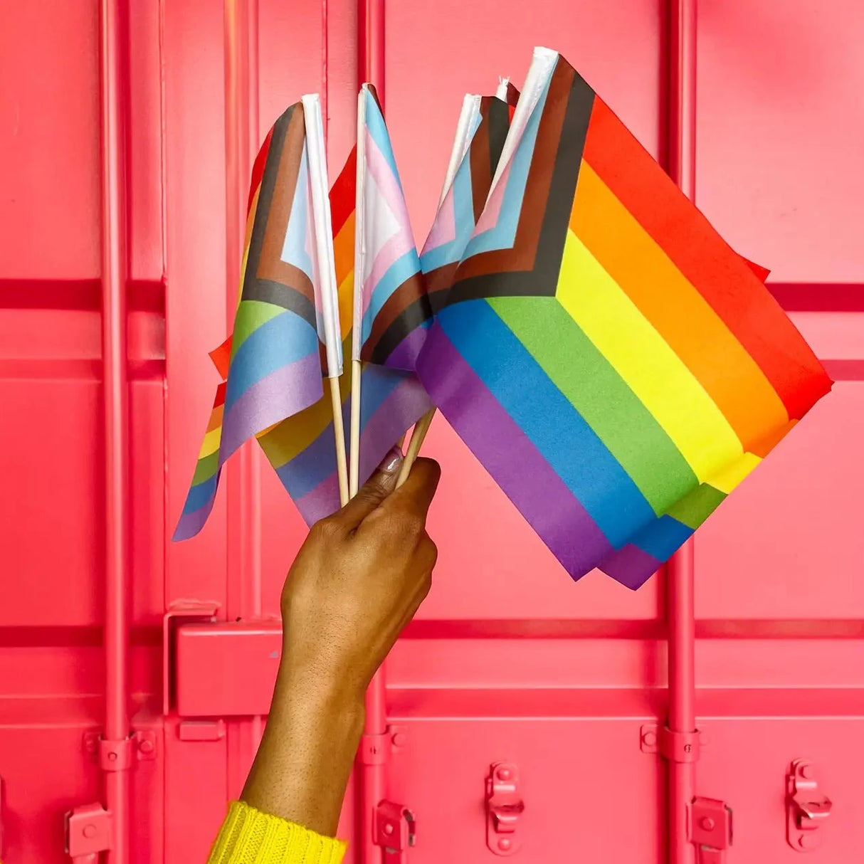 Hand holding several Mini Pride Flag! progress flags with wooden dowels against a bright pink background.