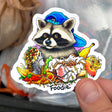 Hand holding Foodie Raccoon with Garbage Sticker showing a raccoon in a blue hat surrounded by colorful food and trash.