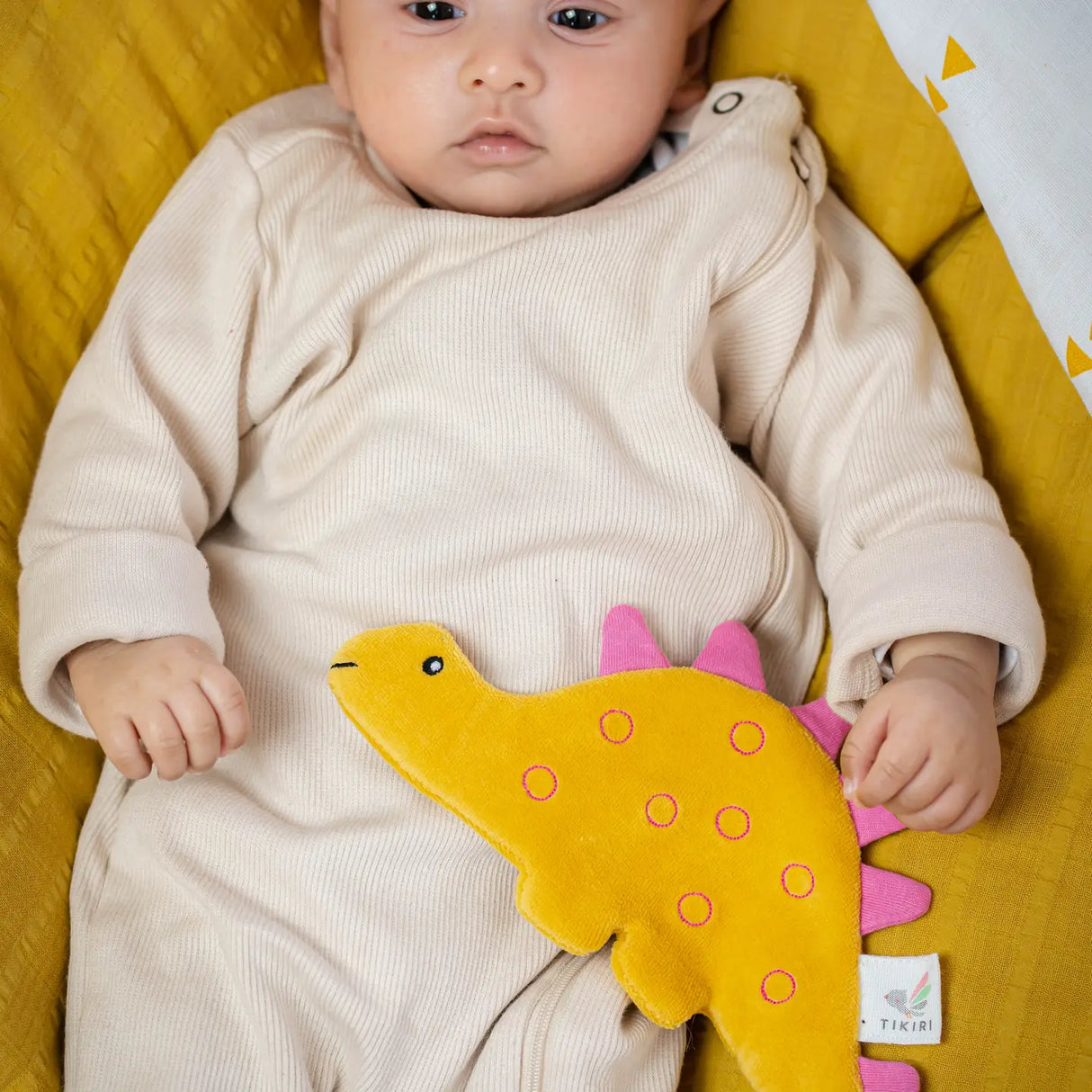Organic Crinkle Fabric | Stego yellow and pink stegosaurus toy held by baby in beige outfit on yellow blanket.