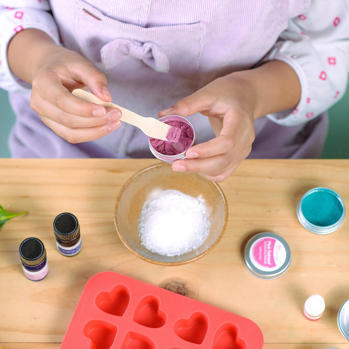 Child scooping purple powder to mix with ingredients in DIY Bath Bombs Kit on wooden table with mold and oils.