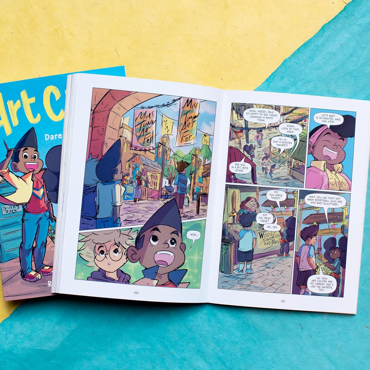 Open pages of the Art Club graphic novel showing colorful comic illustrations featuring young characters in a vibrant city setting.