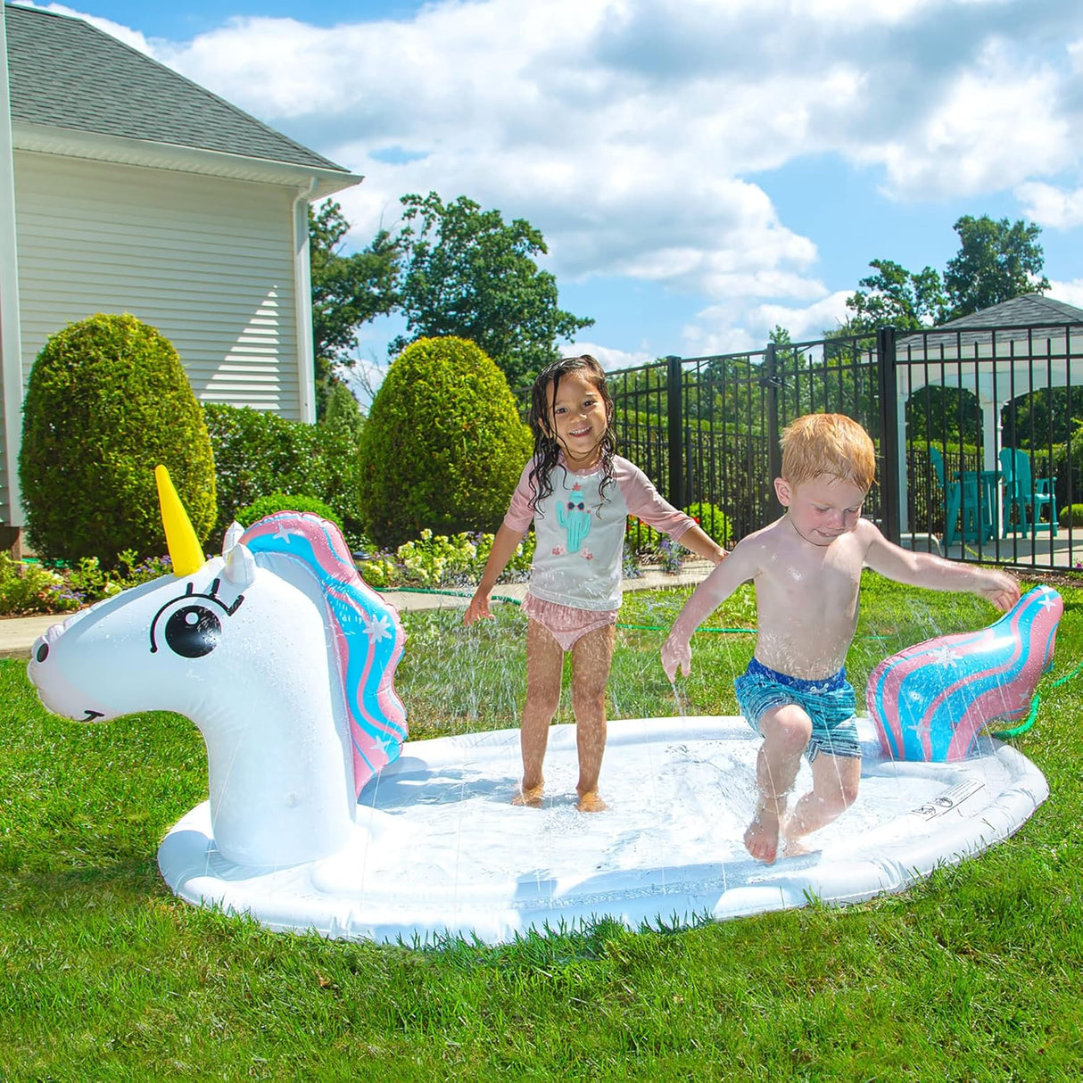 Two children playing and splashing on the Splash Pad Sprinkler Unicorn inflatable water toy outdoors on grass.