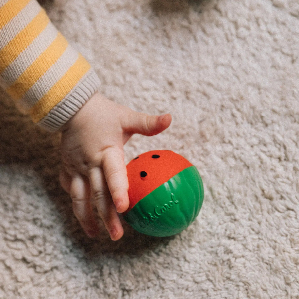 Baby hand holding a textured red and green sensory ball designed to soothe teething and promote motor skills.