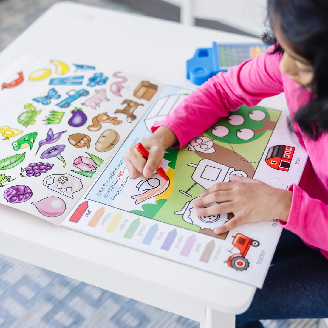 Child coloring a farm scene with crayons in the Colors & Shapes Activity Pad on a white table.