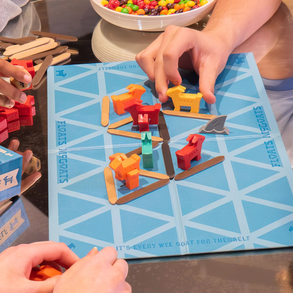 Hands playing the strategic game Floats McGoats with colorful goat pieces and wooden sticks on a blue game board.