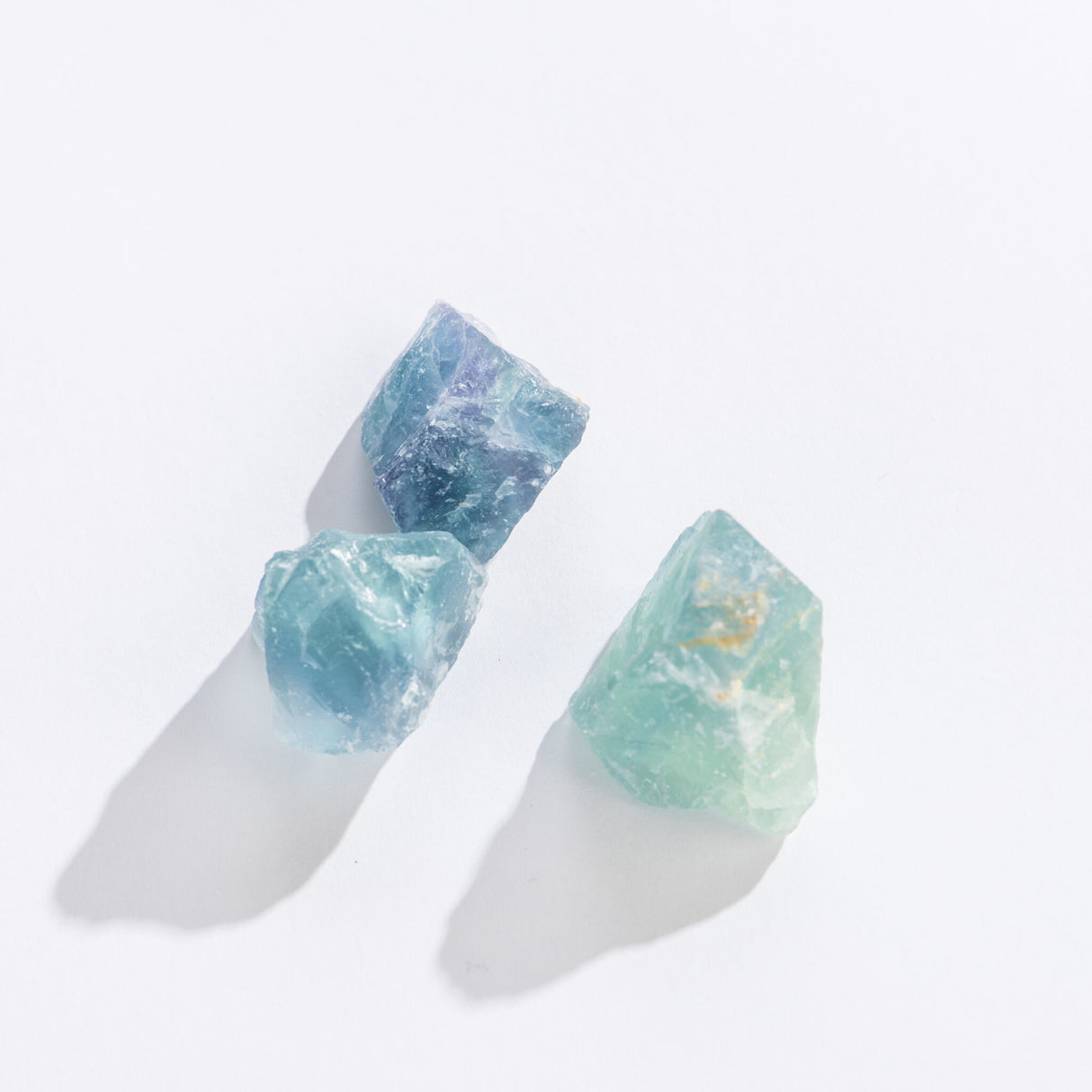 Three rough green fluorite stones in varying shades on a white surface casting soft shadows