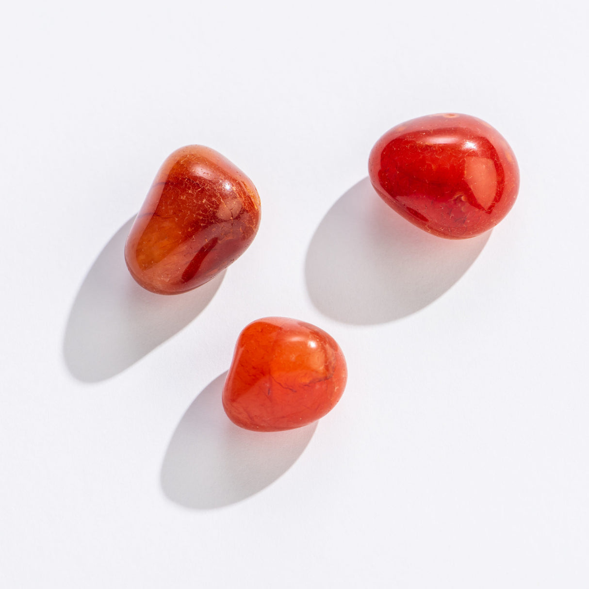 Rock | Tumbled Carnelian Crystal stones in varying orange-red hues on a white background, promoting creativity and healing.