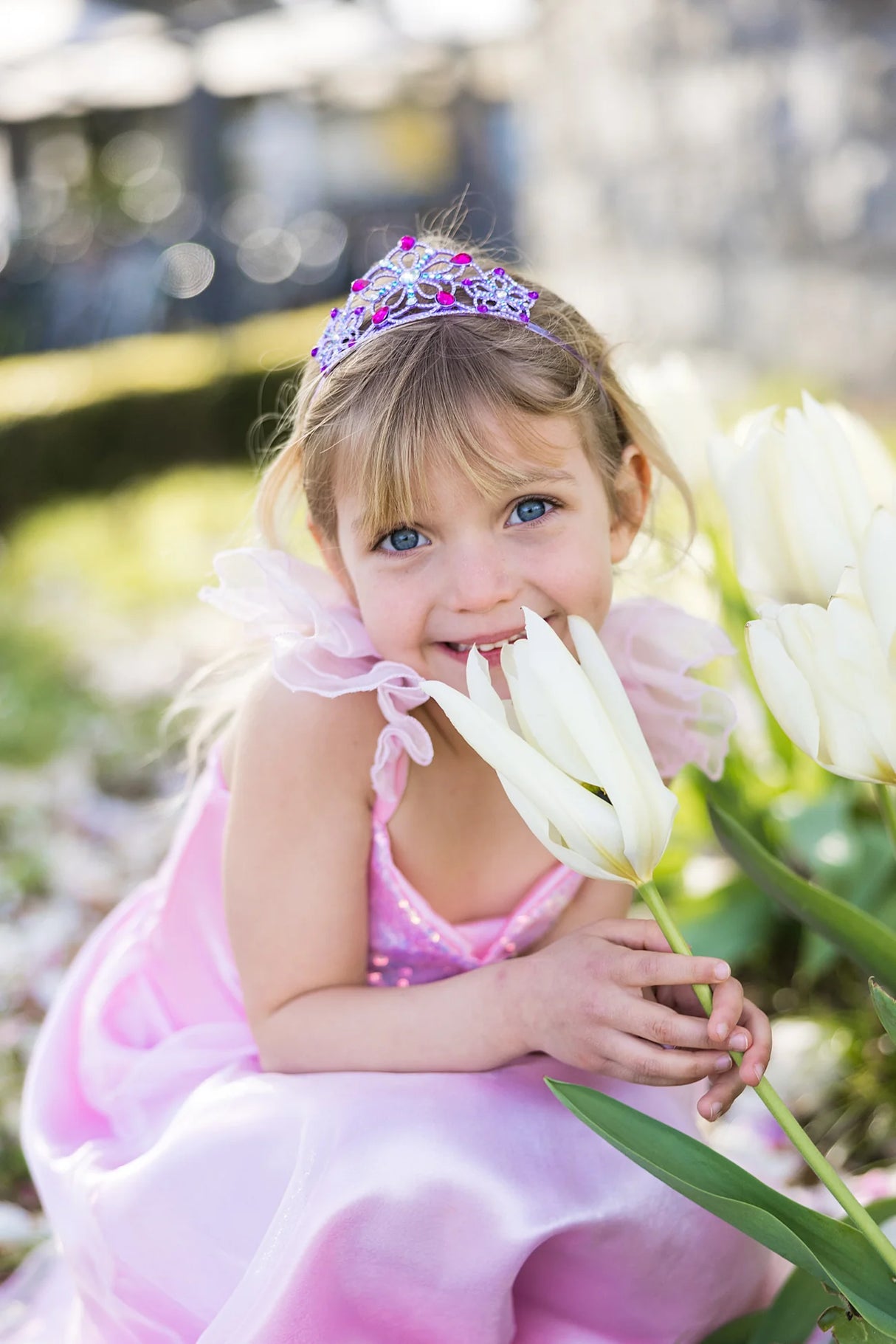 Little girl wearing Be Jewelled Tiara with lilac jewels, holding white tulips, dressed in a pink princess outfit outdoors.