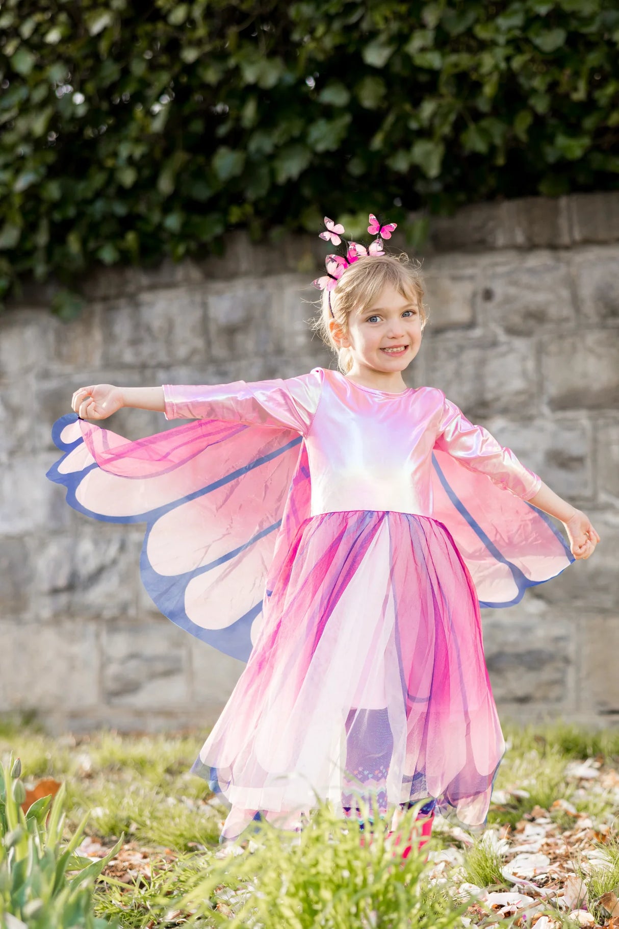 Girl wearing Butterfly Twirl Dress & Wings | Pink with attached butterfly wings and matching headband outside.