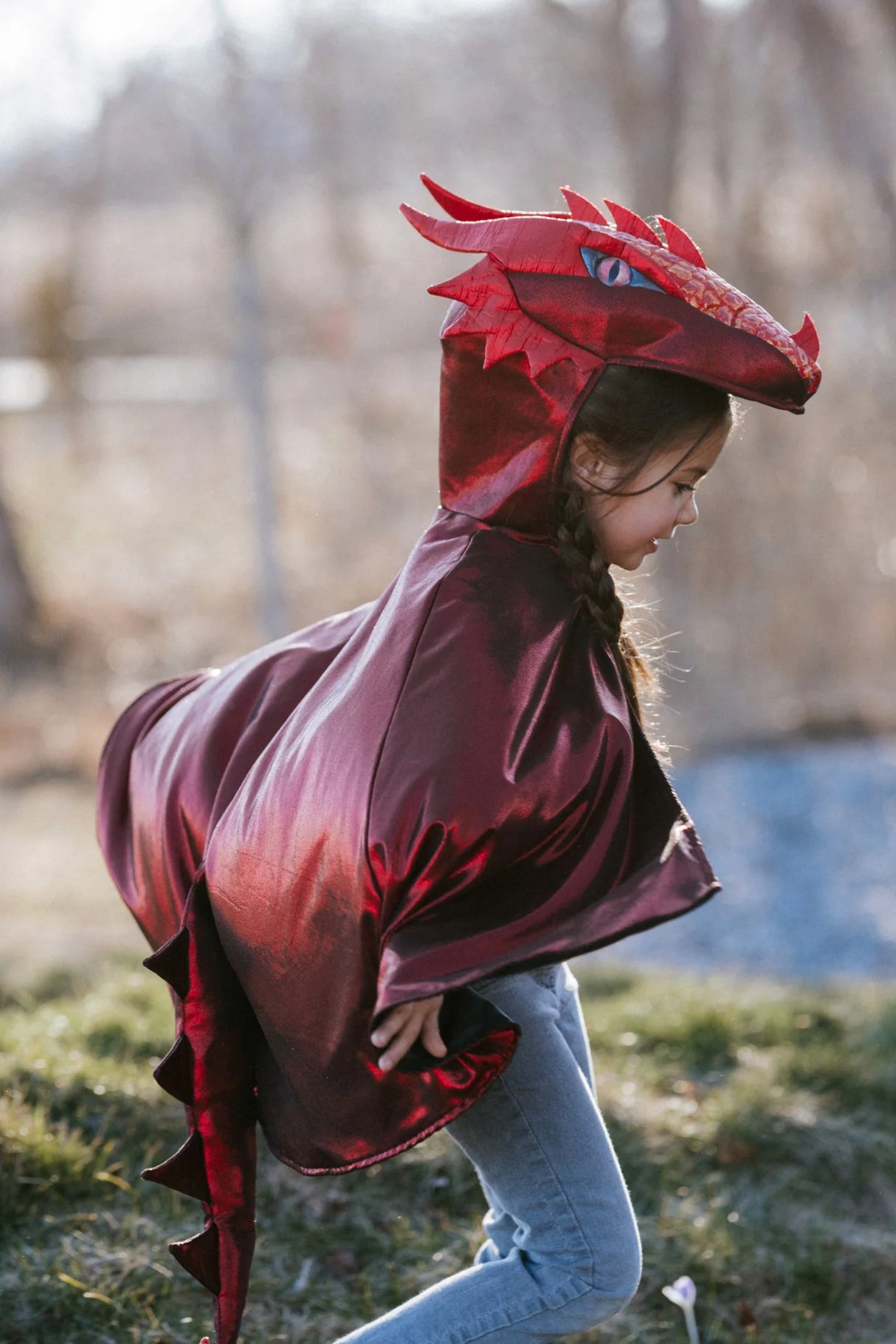 Young child wearing the Ruby the Metallic Dragon Cape Size 5-6 in shimmering red with a 3D dragon head hood outdoors.