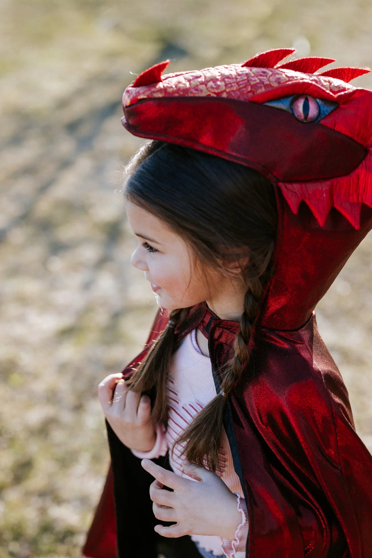 Child wearing Ruby the Metallic Dragon Cape size 5-6 with shiny red dragon hood and scale details outdoors.