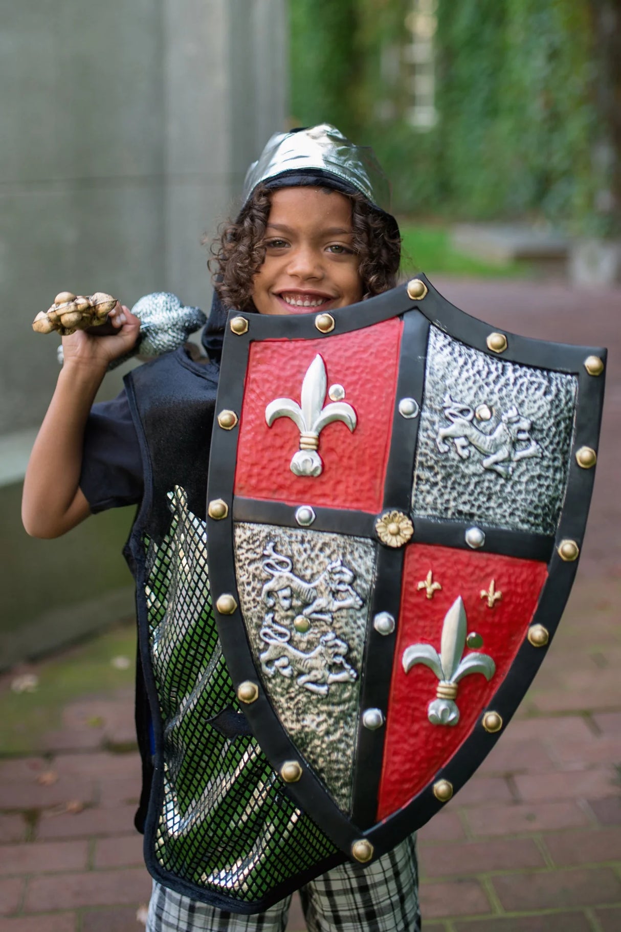 Child dressed as a knight holding the Knight Shield with red, gold, and silver lion and fleur de lis patterns.